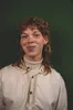 Een medium close-up studioportret van Maxime met een vriendelijke glimlach en rood, krullend haar in een mullet. Maxime draagt een crèmekleurig overhemd over een witte coltrui en een subtiele neuspiercing, gefotografeerd tegen een egale, donkergroene achtergrond.