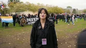 Een vrouw met een zwarte jas en zwart en een perskaart om haar hals kijkt in de camera. Op de achtergrond is een protest gaande met Nederlandse vlaggen en 'Defend the Netherlands'.