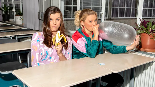 De Sekszusjes zitten in een klaslokaal met een opgeblazen condoom en een banaan.