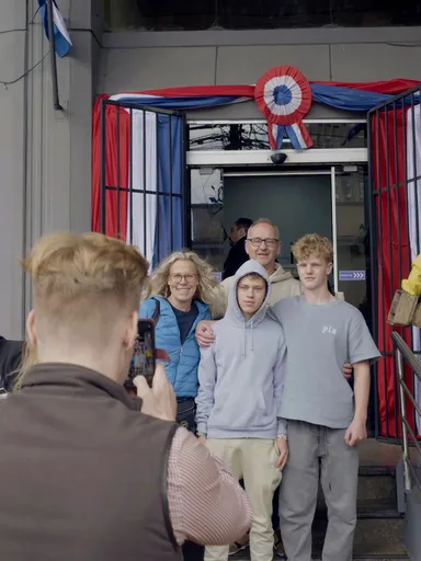 Een wit, Nederlands gezin, bestaande uit een vader, een moeder en twee zoons, poseren voor het gebouw van de migratiedienst in Paraguay, terwijl een jongeman een foto maakt met zijn smartphone.