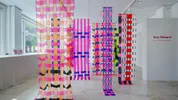 Een lichte expositieruimte met lange, transparante textielbanen die van het plafond hangen. De stoffen hebben geometrische patronen in felle kleuren zoals roze, oranje, paars, blauw en rood. Op de rechterwand staat ‘Amy Tidmarsh – Woven Patchwork: Hybrid Making’.