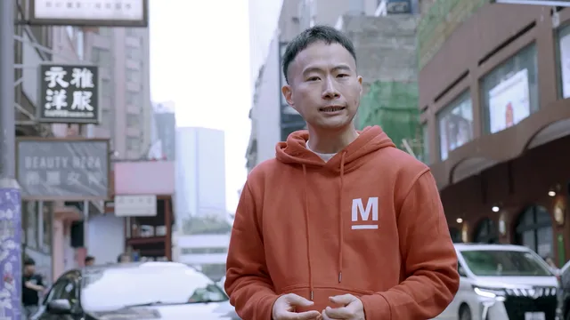 Metropolis-reporter Chang heeft een rode Metropolis-trui en en staat in een Chinese straat. Hij kijkt de camera in.
