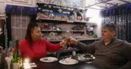 Een vrouw links aan tafel en man rechts aan tafel proosten bij hun avondeten.