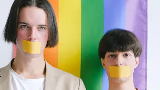 Twee personen die de camera in kijken met een stuk tape over hun mond geplakt. Op de achtergrond een regenboogvlag.
