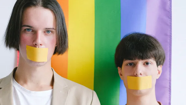 Twee personen die de camera in kijken met een stuk tape over hun mond geplakt. Op de achtergrond een regenboogvlag.