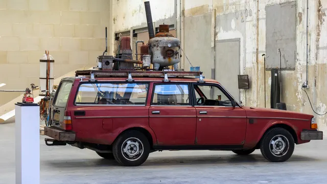 Een oude, donkerrode stationwagen staat in een grote industriële expositieruimte met verweerde muren. Op het dak is een installatie van roestige metalen ketels, buizen en andere apparaten bevestigd. Links naast de auto staat een klein object op een witte sokkel.