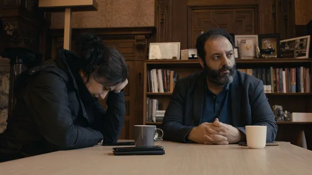 Vrouw (l) en man zitten samen aan een tafel. Achter hen een boekenkast. De vrouw zit met haar hoofd gebogen.