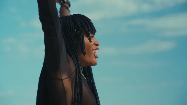 Een jonge vrouw met dreads en piercings door haar neus en lippen is en profiel te zien terwijl ze haar armen in de lucht houdt en lacht.