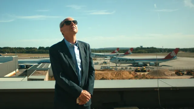 Een witte oudere man in donker pak en zonnebril staat op een terras bij een luchthaven en kijkt omhoog; achter hem staan meerdere vrachtvliegtuigen op het platform onder een heldere blauwe lucht.