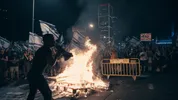 Protestdemonstratie, pallets staan in vuur en een man spreekt een groep demonstranten toe met een megafoon.
