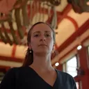 Isabella Clegg staat in een museum onder een groot hangend walvisskelelet. Ze draagt een donker shirt en kijkt ernstig in de camera; de rode staalconstructie van het gebouw is zichtbaar op de achtergrond.