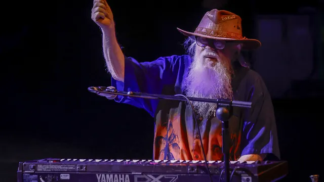 Een man met lange grijze baard met een hoed op zit achter zijn keyboard. Hij draagt een kleurrijk hawaishirt