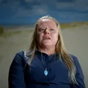 Annemarie van den Berg zit voor een achtergrond met een geschilderd duinlandschap. Ze draagt een donkerblauw overhemd en een blauw stenen hanger, en spreekt terwijl ze omhoogkijkt.