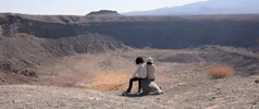 Twee personen zitten op een rots en kijken uit over een grote, droge krater in een bergachtig woestijnlandschap.