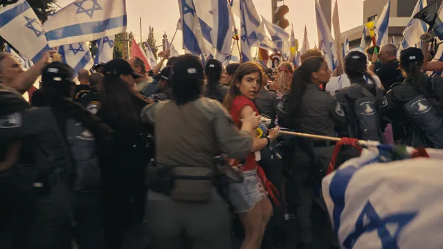 Nava Rozolio,een van de protestleiders. Er is een demonstratie met Israëlische vlaggen en politie op een drukke straat.