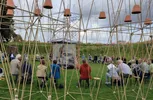 Een groep voornamelijk witte mensen zit in een grote kring op stoelen in een grasveld. Ze worden omringd door een hoge constructie van bamboestokken en touwen, waaraan omgekeerde terracotta bloempotten hangen. In het midden staat een cilindervormig object met doorzichtige wanden en een blauw dak. De lucht is bewolkt.