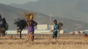 Ethiopische vrouw en jong meisje lopen door een droog veld met fabrieksgebouwen en bergen op de achtergrond, gevolgd door een hond.