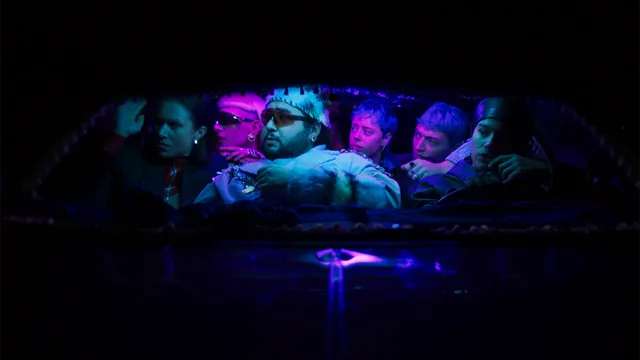 donkere bioscoopzaal met op het scherm een rij mensen die in blauw en paars belicht worden. Ze kijken allemaal naar links en dragen hippe zonnenbrillen.