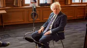 Boris Johnson zit op een stoel in een vergaderzaal. Voor hem staan meerdere microfoons opgesteld voor een interviewopname.