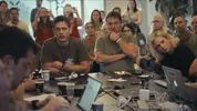 Eyal Naveh zit aan een tafel met meerdere mensen. Om hen heen staan ook veel mensen.