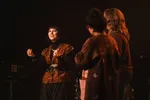 Foto van een vrouw op een podium in een donkere ruimte. Ze draagt een zwart colshirt, een vest met een paisley-patroon en een brede zwarte riem. Ze kijkt glimlachend omhoog en maakt handgebaren terwijl ze tegen twee mensen op de voorgrond praat, die op de rug worden gezien. Op de achtergrond zijn vaag een keyboard en drumstel zichtbaar.