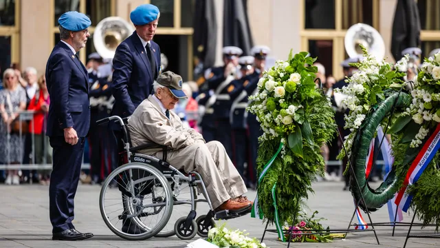 En oude man in een rolstoel, vergezeld door twee militairen, bij een kranslegging bij de herdenking van het bombardement van 14 mei 1940 op Rotterdam.