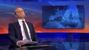 Nieuwslezer in pak zit aan een desk in een televisiestudio met een kaart van Europa op een scherm achter hem, kijkt serieus naar de camera terwijl hij papieren vasthoudt.