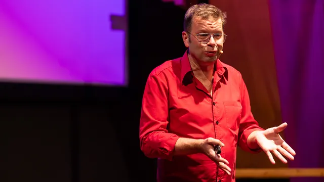 Een man met bril in rood overhemd spreekt op een podium