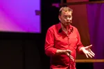 Een man met bril in rood overhemd spreekt op een podium