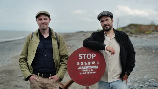 Twee mannen staan op een kiezelstrand bij een rood stopbord. Links een man in groene jas, rechts een man in zwarte jas.