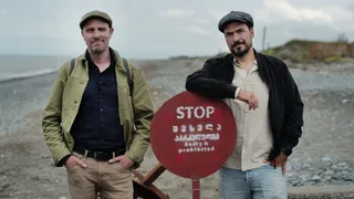 Twee mannen staan op een kiezelstrand bij een rood stopbord. Links een man in groene jas, rechts een man in zwarte jas.