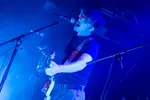 Een actiefoto van een gitarist met een bril die gepassioneerd zingt in een microfoon tijdens een optreden. Hij draagt een donker T-shirt en een gitaarband met een kleurrijk patroon. Het podium is fel verlicht met een diepblauwe gloed en op de achtergrond is vaag een tweede muzikant zichtbaar.