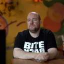 Alex Romijn zit aan een tafel in een kleurrijk café met een geschilderde muur op de achtergrond. Hij draagt een zwart T-shirt met de tekst "Bite Back" en kijkt recht in de camera.