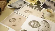Er liggen oude portretten en foto's op een tafel. Op tafel staat ook een kop koffie.