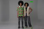 Twee zwarte personen met donker krullend haar staan naast elkaar tegen een grijze achtergrond. De linkerpersoon heeft een grote afro en draagt een kleurrijk shirt met een groen-roze patroon en een gescheurde spijkerbroek. De rechterpersoon draagt een beige mouwloos vest, een zwarte crop top en een wijde donkergrijze broek. Om de rechterpersoon staat een groen kader met bovenaan het label ‘PERSON’.