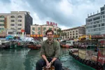 Een witte man met donkerbruin haar en een bril zit op een krukje aan het water, met een legergroen overhemd aan. Achter hem liggen kleurrijke vissersboten in een haven, omringd door gebouwen met Chinese opschriften en een Chinese vlag. De lucht is bewolkt.