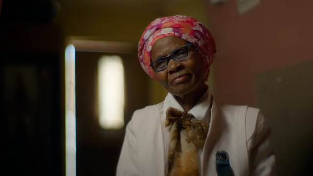 Een vrouw met een donkere huidskleur zit in een kamer, ze heeft een oranje doek met print om haar hoofd, een zwarte bril op en een chique wit jasje aan met een licht bruine sjaal.