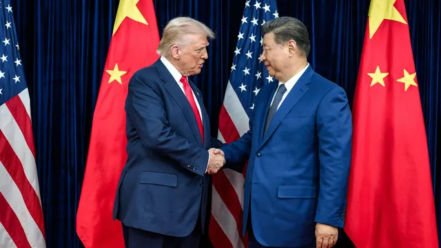 Een witte man met blond haar en rode stropdas schudt de hand van een Oost-Aziatische man met lichtbruine huid en grijszwart haar. Achter hen staan Amerikaanse en Chinese vlaggen.
