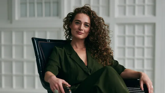 Brankele Frank zit op een zwarte stoel met armleuningen, ze kijkt de camera in en heeft een donkergroen pak aan.