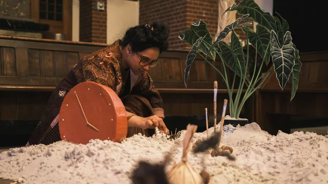 Een persoon met een bril en een patroonjasje knielt neer in een kerk, gericht op een installatie. De persoon reikt naar de installatie, die bestaat uit een zanderige of suikerachtige heuvel, een grote rode klokachtige structuur, een pot met een grote plant en diverse borstels en stokken. Een onscherp object bevindt zich op de voorgrond. De achtergrond toont houten banken en een bakstenen muur, met een standbeeld van een religieuze figuur op een voetstuk aan de rechterkant.