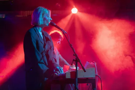 Een sfeervol beeld van een zangeres met kort blond haar die tegelijkertijd een synthesizer bespeelt op een podium. Ze is gehuld in een donkere blazer en staat voor een microfoon, terwijl ze baadt in een fel rood en blauw tegenlicht met rook op de achtergrond. Naast haar is vaag een ander bandlid achter apparatuur zichtbaar.