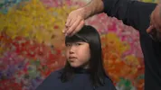 Bo zit in de kappersstoel, met een schuin oog kijkt ze richting de camera. Marko knipt haar pony.