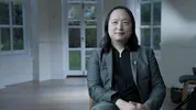 Een man met halflang haar zit in een lichte ruimte met hoge ramen en openslaande deuren op de achtergrond. De persoon draagt een grijs pak en kijkt ontspannen in de camera.