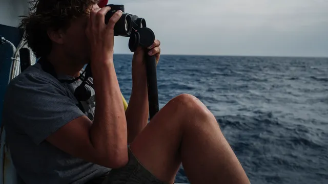 Rob Timmerman zit op het dek en kijkt met een verrekijker naar de zee