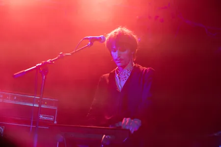 Een sfeervol beeld van een muzikant met warrig haar en een opvallende halsketting, die geconcentreerd op een keyboard speelt. De persoon draagt een donker overhemd en bevindt zich op een podium dat baadt in een warme, rode gloed met een lichte mist. Links in beeld staat een microfoon op een standaard en een Ampeg-versterker.