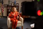 Een vrouw in een rood pak zit op een stoel tijdens een interview in een kantoorruimte. Ze maakt een handgebaar terwijl ze spreekt.