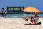 In de branding op het strand van Tel Aviv staat een protestbord voor het thuisbrengen van de Israëlische gijzelaars. In het zand daaromheen lopen en liggen badgasten te genieten van de zon.