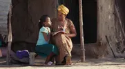 Moeder leest een boek met haar dochter op een houten bankje voor een traditionele lemen hut in een dorpsomgeving.