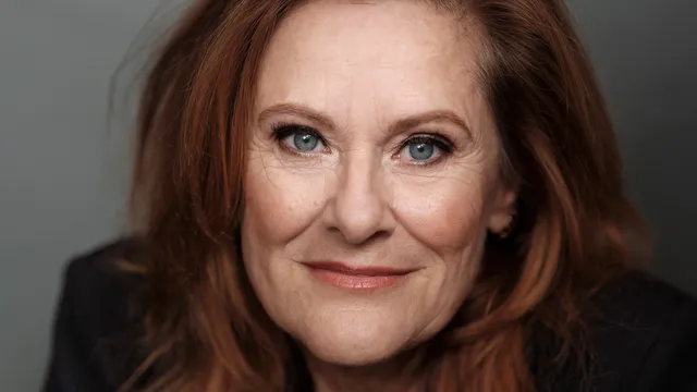 Close-up portret van Lotje IJzermans. Een vrouw met roodbruin haar, blauwe ogen en een subtiele glimlach, gefotografeerd tegen een neutrale grijze achtergrond.