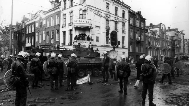 Een straathoek in Amsterdam. Op de achtergrond een groep krakers op een balkon met spandoeken. Op de voorgrond in de straat een tank en agenten in zwarte kleding met helmen en schilden.